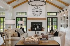a neutral barn living room with white planked walls and a ceiling, wooden beams, a fireplace, neutral seating furniture, coffee tables and a metal chandelier