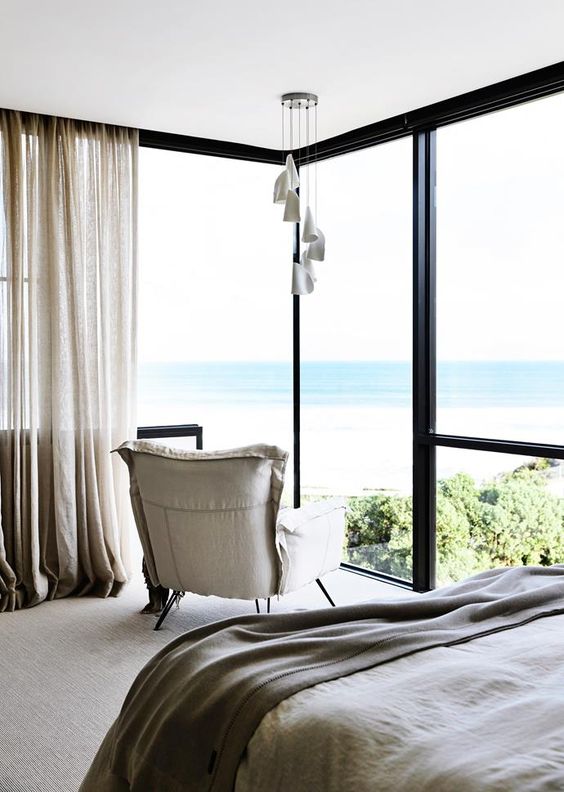 a neutral bedroom with a floor to ceiling corner window that provides gorgeous beach and sea views and a chair to enjoy them