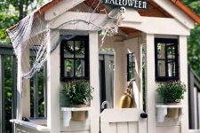 a neutral kids’ playhouse with a roof, with black window frames, potted plants and blooms styled for Halloween