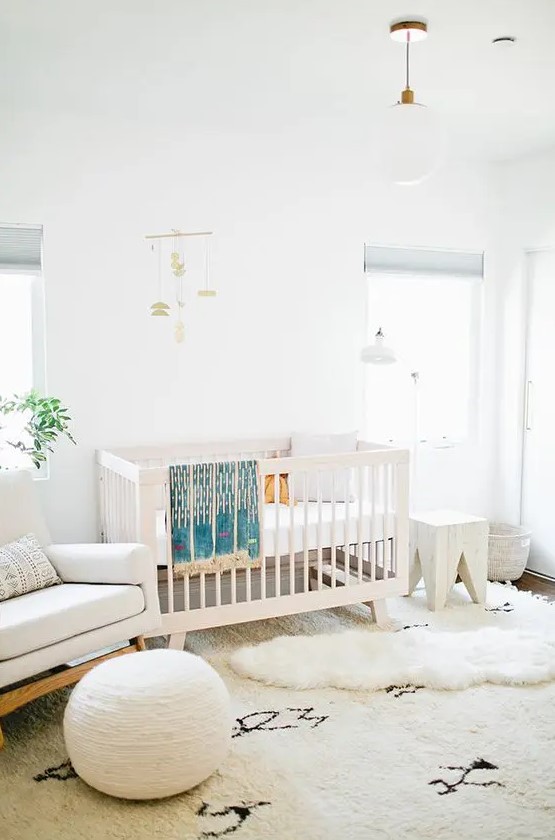 a neutral mid century modern meets boho nursery with a white crib, a creamy rocker chair, a round pouf, a white stool, layered rugs