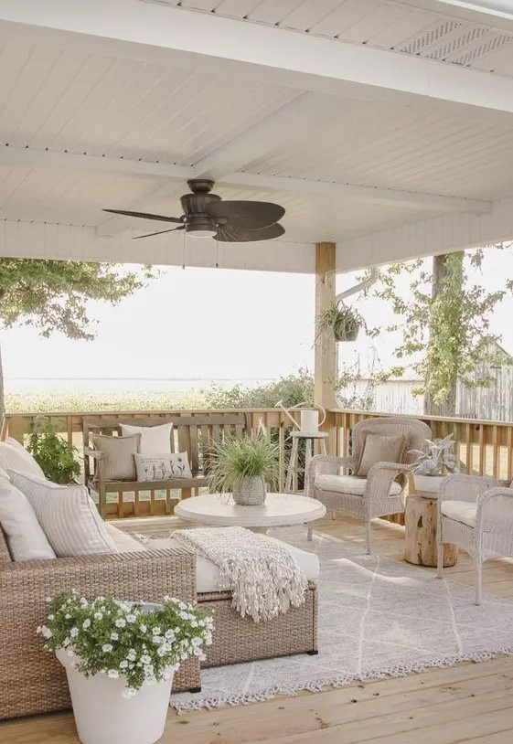 a neutral rustic seaside terrace with light colored wicker furniture, white upholstery and textiles and potted blooms and greenery