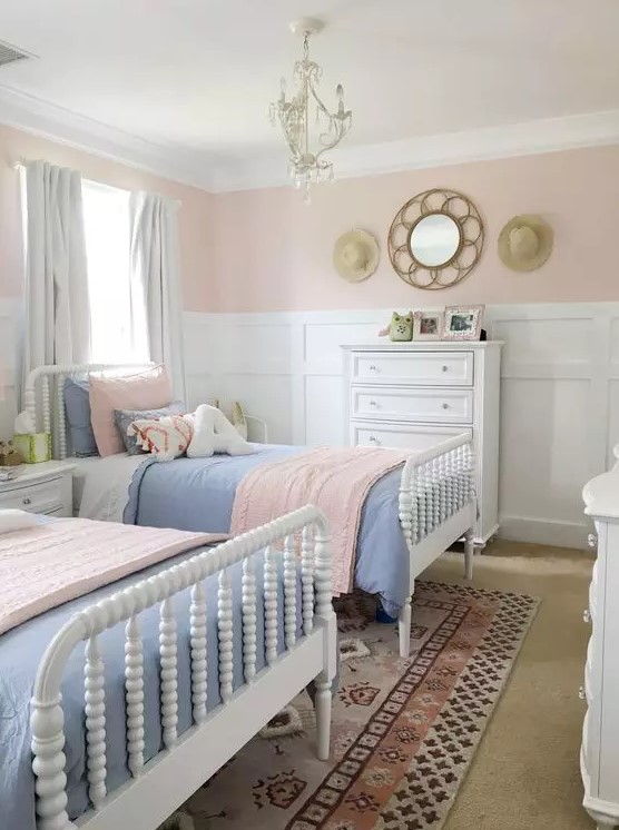 a pastel farmhouse shared girls' bedroom with white paneling on the walls, white vintage beds, pink and blue bedding and a vintage chandelier