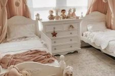 a pastel shared girls’ bedroom with a grey accent wall, white vintage beds, pink and white bedding, a white vintage dresser, neutral and pastel toys