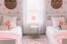 a pretty and bright shared girls’ bedroom with a polka dot accent wall, white farmhouse beds, pink and white bedding, a colorful rug and names