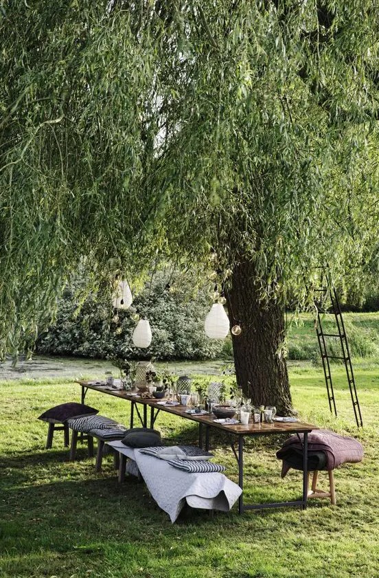 a pretty and cool garden dining zone under the tree, with a long table, benches and a stool, pillows and blankets, pendant lamps