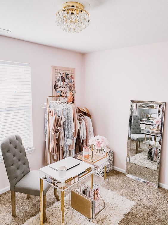 a pretty and delicate glam cloffice with a makeshift closet, a mirror, an acrylic desk and a chest, a grey chair and a glam chandelier
