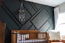 a pretty and small mid-century modern nursery with a black paneled wall, a stained crib, an amber leather chair, neutral textiles, a chandelier and a mobile