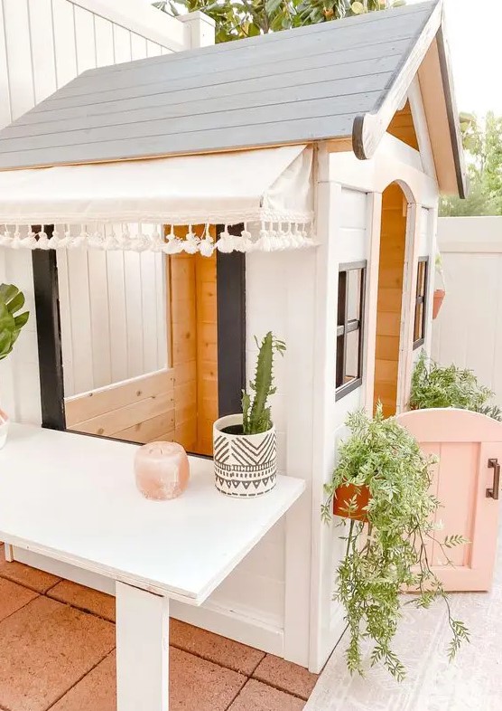 a pretty black and white kids' playhouse with a pink half door, with potted greenery and plants, with a large table next to it
