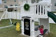 a pretty kids’ playhouse of white planks, with a slide, a black door, a built-in dining space, swings and a greenery wreath