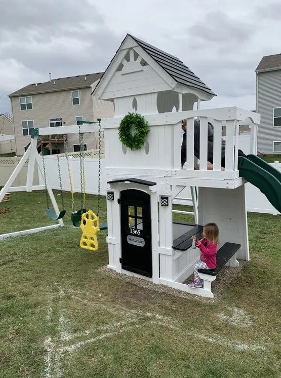 a pretty kids' playhouse of white planks, with a slide, a black door, a built in dining space, swings and a greenery wreath