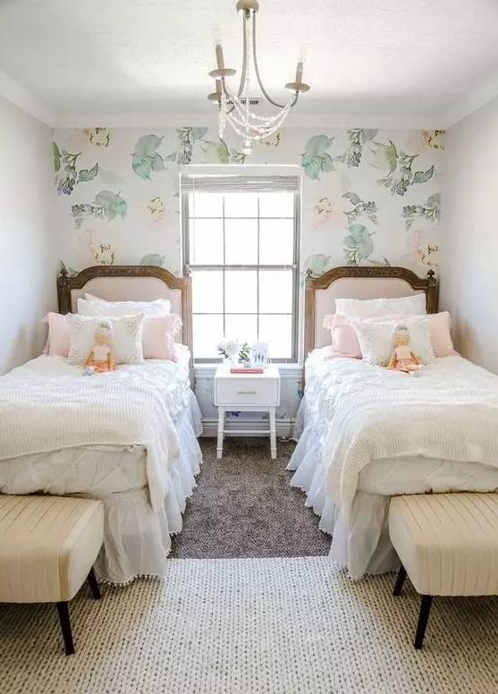 a pretty shared girls' bedroom with a pink floral accent wall, pink upholstered beds, creamy poufs and a vintage chandelier