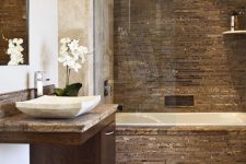 a refined bathroom clad with neutral stone tiles, with a brown accent wall and cladding on the tub, a floating dark-stained vanity with a neutral stone sink