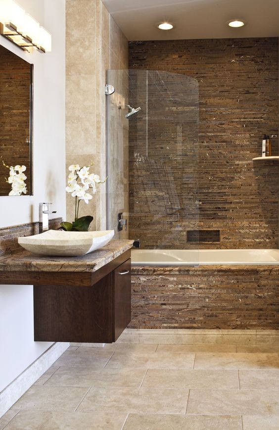 a refined bathroom clad with neutral stone tiles, with a brown accent wall and cladding on the tub, a floating dark stained vanity with a neutral stone sink