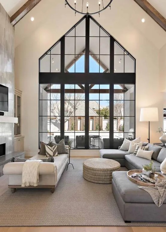 a refined modern living room with double-height windows, a fireplace clad with tiles, a grey sectional, white daybed and a round pouf
