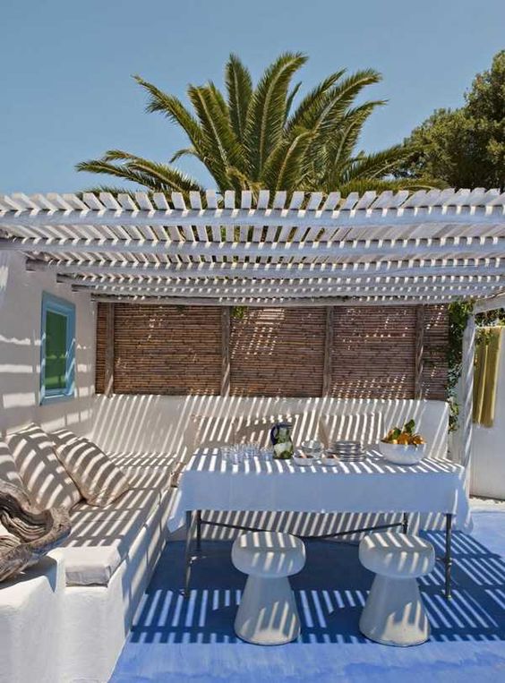 a relaxed coastal terrace with a built-in corner sofa, a white table and stools, a roof and a blue rug is all you need to have a great meal with a view