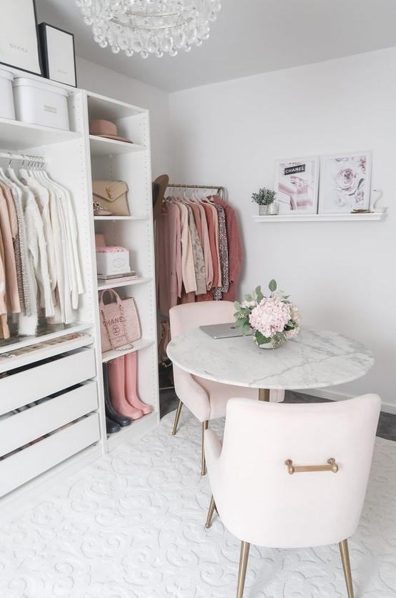 a romantic cloffice with a white makeshift closet, a crystal chandelier, some artworks, blush chairs and a marble table