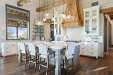 a rustic barn kitchen with light-stained wooden beams and a matching hood, a white kitchen island with tall stools, white cabinets and a cluster of pendant lamps