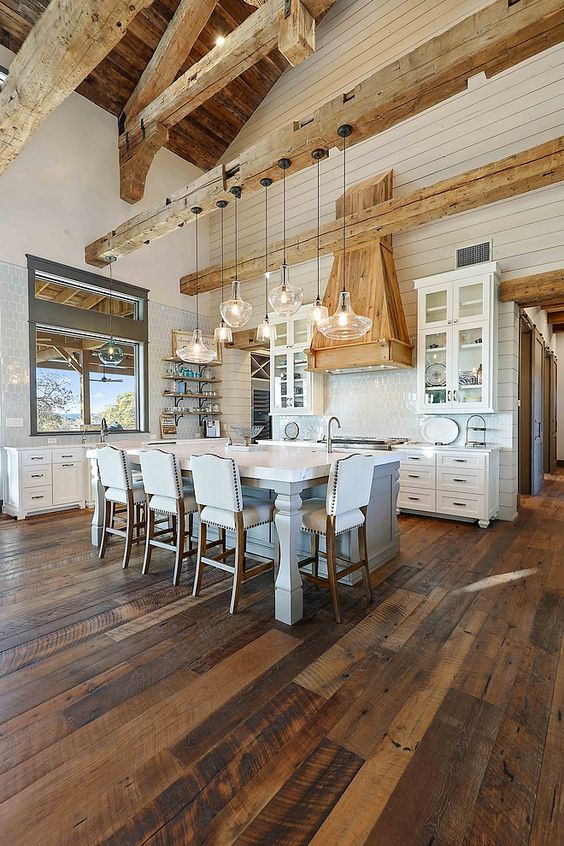 a rustic barn kitchen with light-stained wooden beams and a matching hood, a white kitchen island with tall stools, white cabinets and a cluster of pendant lamps
