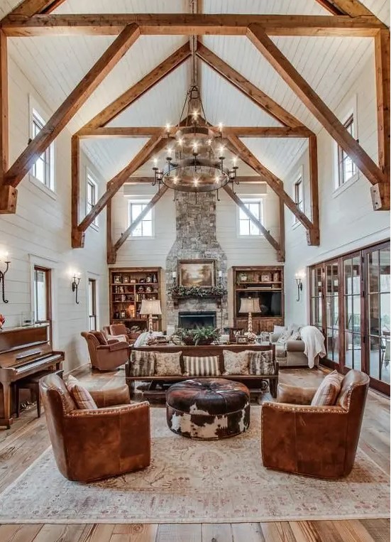 a rustic barn living room with wooden beams and white planked walls and a ceiling, leather furniture, a metal tiered chandelier and neutral textiles