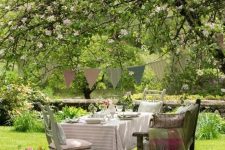a shabby chic dining area under a blooming tree, with a table, chairs and a bench plus pastel textiles