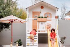 a small and lovely kids’ playhouse in white and stained wood, with climbing part, a slide and potted greenery and a dining space with an umbrella