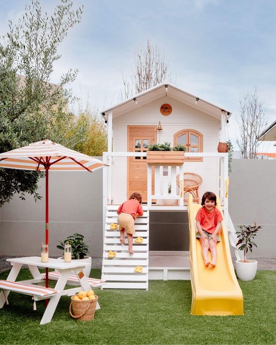 a small and lovely kids' playhouse in white and stained wood, with climbing part, a slide and potted greenery and a dining space with an umbrella