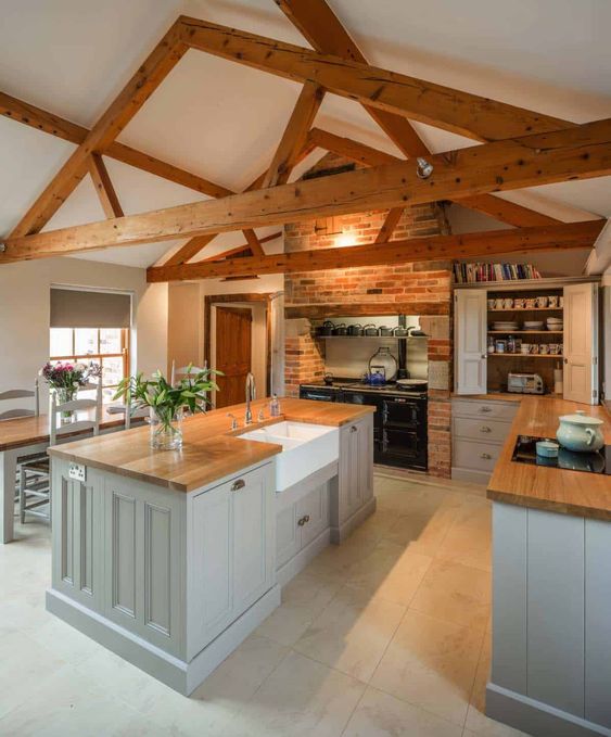 a small barn kitchen with wooden beams, grey shaker style cabinetry, butcherblock countertops, a vintage hearth and some lights