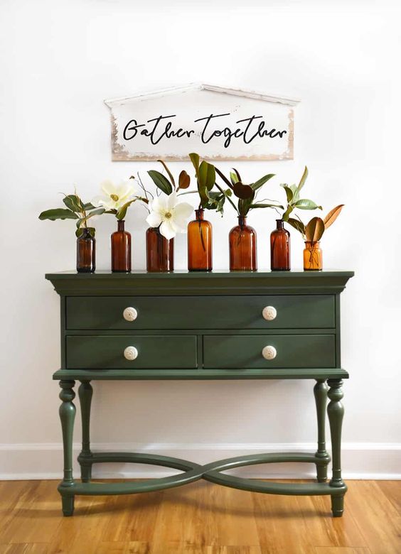 a small green vintage dresser with an arrangement of apothecary bottles with neutral blooms and a sign over it for styling a rustic space