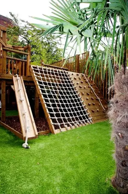 a small outdoor playground with green lawn, ladders, nets, a small tree house and a sand box under it