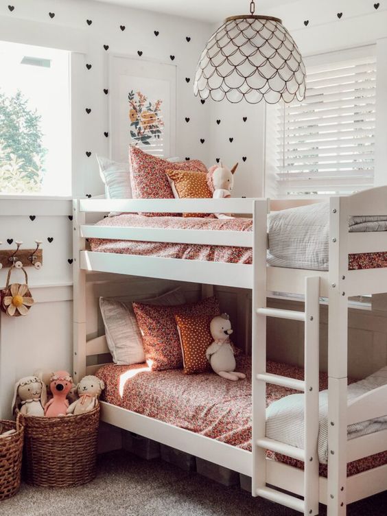 a small shared bedroom with a white bunk bed with floral bedding, baskets with toys and some artwork, a pendant lamp