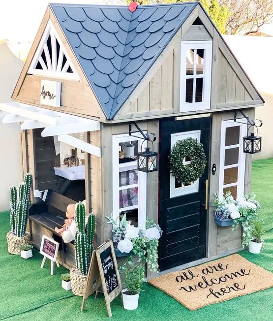 a stylish farmhouse playhouse with a black door, white window frames, a built in bench, potted blooms and cacti