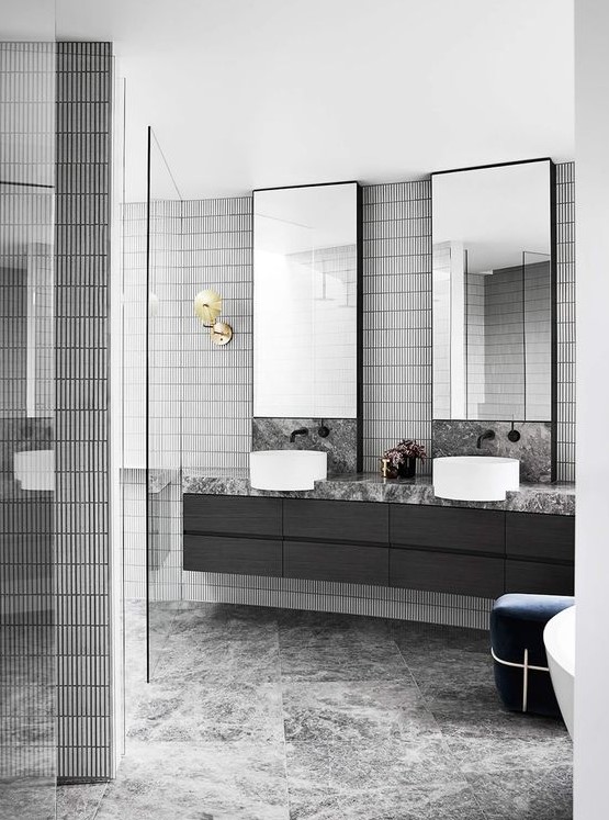 a stylish grey bathroom done with various kinds of tiles, a black floating vanity, two mirrors and sinks and a navy ottoman