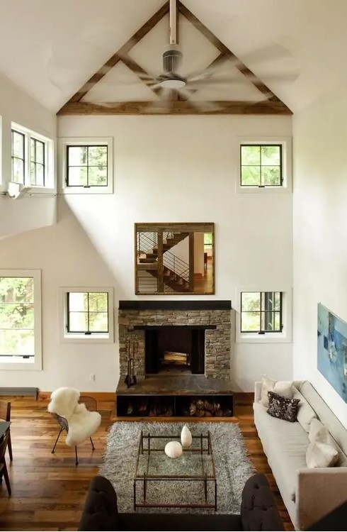 a stylish neutral barn living room with a double-height ceiling, a fireplace clad with stone, neutral seating furniture, a glass table and faux fur is cool