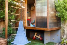 a super cool kids’ playhouse in modern style, with glass walls and a pendant chair, a climbing wall and some bright furniture inside