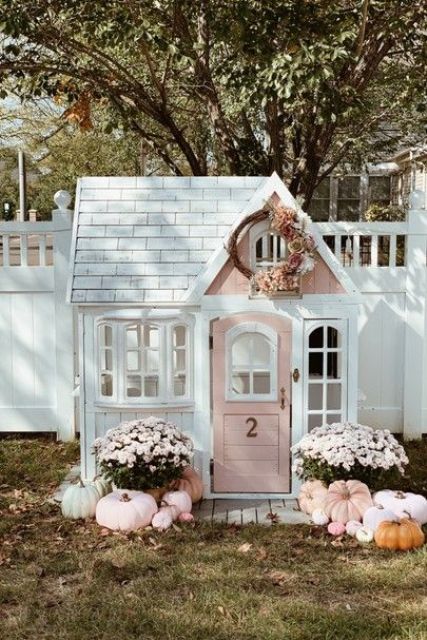 a super cute white and pink kids' playhouse with windows, potted blooms and pumpkins outside and a floral wreath is wow