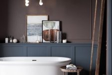a super refined moody bathroom with chocolate brown walls, grey paneling, a chic tub on a marble platform and a floor mirror