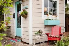 a tan kids’ house clad with siding, with a turquoise door and a window with a turquoise planter and blooms looks like a real personal house