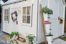 a white farmhouse playhouse with lights, greenery and blooms, a ladder with plants in buckets and a wreath is super cute