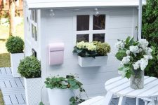 a white kids’ playhouse of planks, with potted blooms and greenery and a pink postbox plus a white wooden dining set in front of it