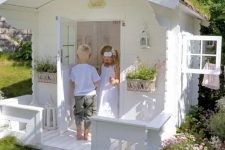 a white kids’ playhouse with carved touches, a white interior and potted blooms looks like a real one