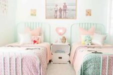 a white shared girls’ bedroom with mint beds and pink and mint bedding, a gallery wall, a white nightstand, a gold chair with bright pillows