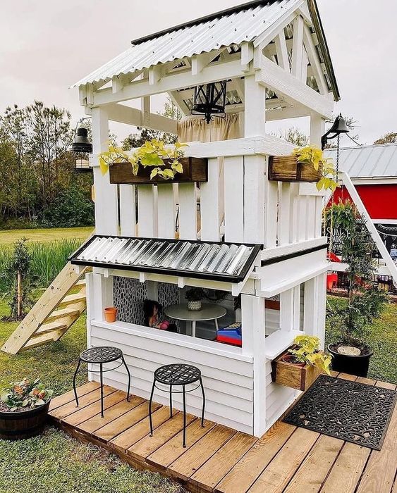 a white two storey kids' playhouse with a little dining space and sleeping one, with stools and a large ladder