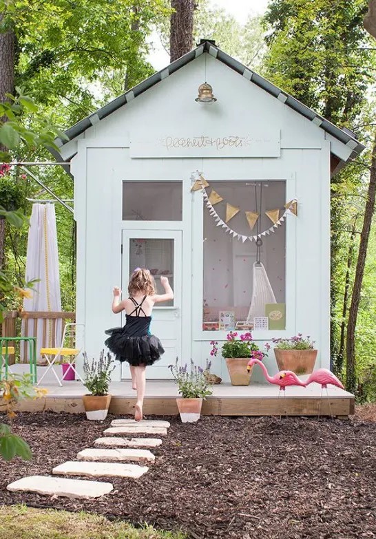 a whitewashed kid's playhouse with a large window, some cool furniture inside, outdoor furniture and potted blooms plus a flamingo