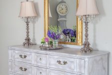 a whitewashed vintage dresser with vintage handles, with vintage table lamps, a mirror in a gilded frame and bold blooms
