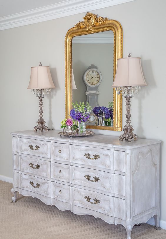 a whitewashed vintage dresser with vintage handles, with vintage table lamps, a mirror in a gilded frame and bold blooms