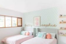 an adorable modern girls’ bedroom with a mint printed accent wall, grey upholstered beds, pink and pastel bedding, jute rugs, wooden shelves and names on the wall