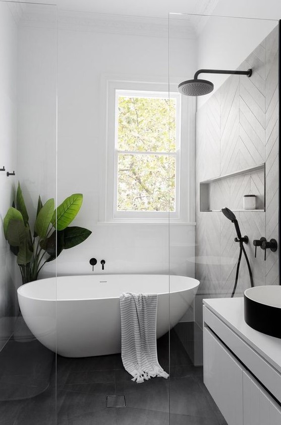 an airy Scandinavian bathroom with grey and white tiles, a floating white vanity, a black sink, a free standing tub and a statement plant