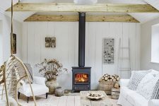 an airy and neutral barn living room with skylights, wooden beams, a dark hearth, white seating furniture, a wooden coffee table and dried blooms