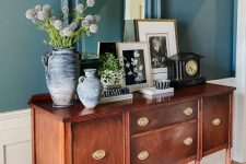 an antique stained dresser with gold knobs, with vases and blooms, artwork, a black clock is a beautiful addition for any space