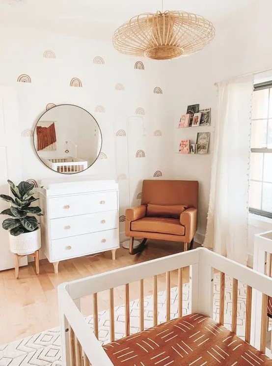 an earthy tone mid century modern nursery with pretty wallpaper, a white dresser, an amber leather chair, a white crib, a wooden pendant lamp and books